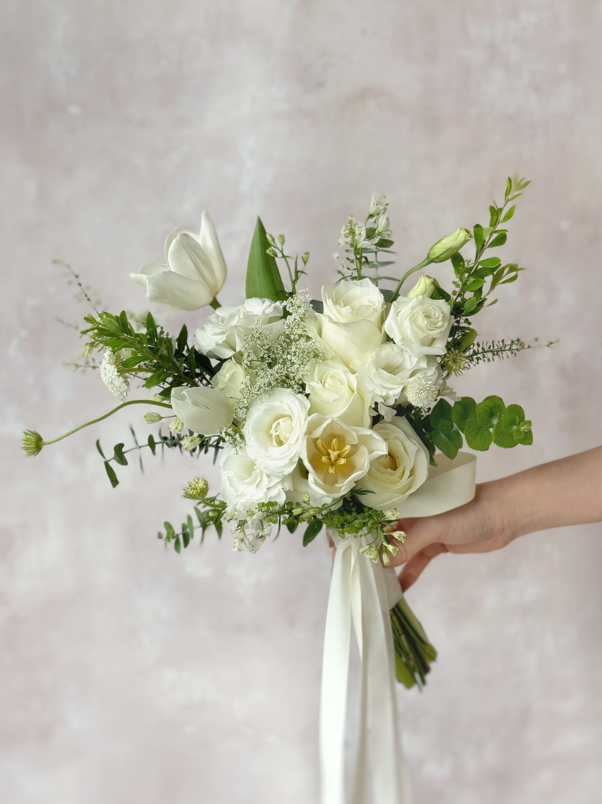 A delicate bridal bouquet featuring white tulips, roses, lace flowers with lush greenery, exuding elegance and natural beauty, held by bride's hand
精致且充满自然气息的白色捧花，适合优雅浪漫的婚礼主题


