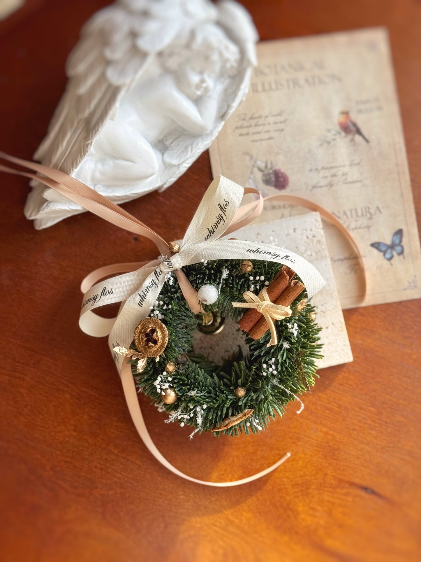 Small decorative woodland-style mini Nobilis fir wreath with earthy accent, cinnamon sticks and ribbons on wooden table.
温馨森系迷你圣诞花环，使用新鲜诺比松、松果和自然材质设计。